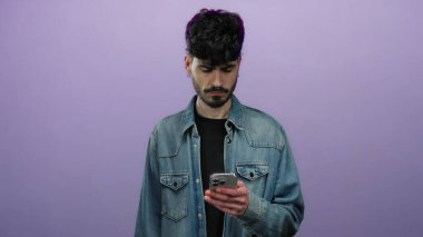 Akıllı telefonlu genç adam, izole edilmiş mor arka plana karşı hayal kırıklığına uğramış görünüyor. Modern dijital hayal kırıklığını, şık bir görünüme ve derin derin düşüncelere sahip bir duruşla ifade ediyor..