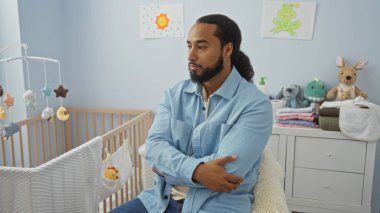 Sakallı genç bir adam bebek beşiğinin yanında rahat bir yatak odasında oturuyor. Oyuncak ve dekorasyonlarla çevrili. Huzurlu bir ev tasviri yapıyor..