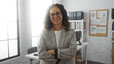 Kollarını kavuşturmuş, kendine güveni tam bir kadın parlak bir ofiste, gözlüklü, ceketli modern bir iş yerinde profesyonelliği ve olgunluğu yansıtan bir şekilde duruyor..