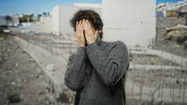 İnşaat alanında yüzünü örten, duygularını ifade eden, gri süveter giyen açık havada duran, berrak mavi gökyüzünün altında bulanık şehir planına sahip genç bir adam..