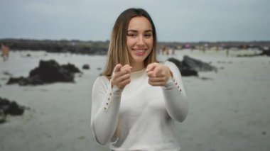 Genç bir kadın gülüyor ve parmaklarıyla deniz kıyısında bulutlu bir gökyüzünün altında kayalık bir kıyı şeridini işaret ediyor..