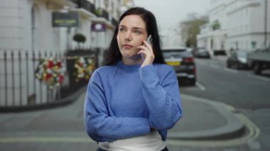 Ciddi bir ifadeyle bir kadın, şehir manzarası ve bulutlu bir günde park halindeki arabalarla çevrili, mavi bir kazak ve koyu bir pantolon giyerek şehir caddesinde duruyor..