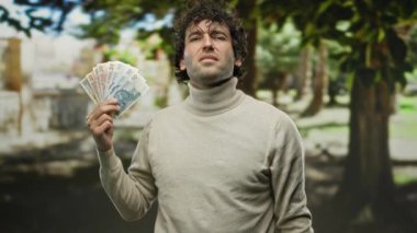 İspanyol bir adam park ortamında zloti banknotlar tutuyor, açık hava yeşilliğinin ortasında kendinden emin bir ifadeyle jestler yapıyor..