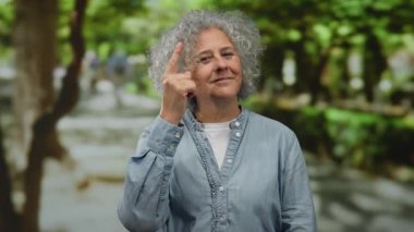 Gri saçlı yaşlı kadın, güneşli bir parkta, rahat bir şekilde el kol hareketi yapıyor, rahat bir kot gömlek giyiyor, yemyeşil ve ağaçlarla çevrili, sakin bir günde..