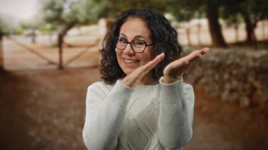 Açık hava parkında kıvırcık saçlı İspanyol kadın şakacı bir jest yapıyor, para harcadığını gösteriyor, doğal ortamda neşeli bir yüz ifadesi olan rahat bir kazak giyiyor..