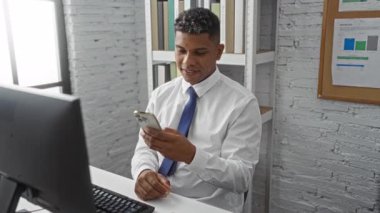 Ofiste akıllı telefon kullanan, gülümseyen, odaklanan, bilgisayarla, belgelerle, profesyonel ortamda masasında oturan, beyaz gömlek ve mavi kravat takan, çalışan, kapalı alanda çalışan genç bir adam.