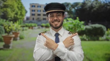 Pilot üniforması ve şapkası içinde sakallı, gülümseyen genç İspanyol adam yeşil bir parkta duruyor. Arka planda bulanık bir bina var..