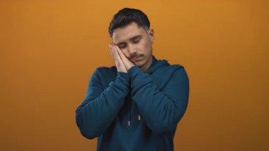 İzole edilmiş turuncu bir arka plana karşı esneyen ve uyuyan genç İspanyol bir adam mavi bir kapüşonlu giyip rahatlama ve huzuru iletiyor.