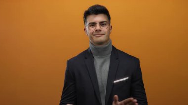 Gözlüklü ve balıkçı yakalı genç adam izole edilmiş turuncu bir arka plana karşı duruyor ve modern profesyonel bir hava yayıyor..