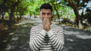 Çizgili gömlekli genç adam yeşillik ve gölgelerle çevrili güneşli bir park ortamında şaşırmış görünüyor..