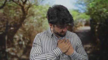 Sakallı genç bir adam parkta göğsünü sıkıyor, muhtemelen yeşil ağaçlarla çevrili kalp rahatsızlığı yaşıyor ve sıkıntılı bir gün ışığı görüyor..