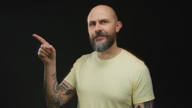 Stüdyoda siyah arkaplanı işaret eden dövmeli ve sakallı bir adam odaklanma ya da karar verme konseptini belirliyor.