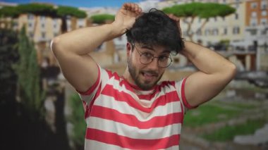 Kırmızı çizgili tişörtlü, sakallı genç İspanyol adam güneşli Roma harabelerinde kulak hareketi yaparak antik mimari ortamında neşeli ve güneşli bir anı yakalıyor..