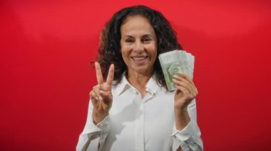 Beyaz gömlekli orta yaşlı bir kadın canlı kırmızı bir duvara karşı Şili pezosu taşıyor. Bu da izole bir ortamda başarı ve finans simgesi..