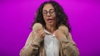 Kadın kürk yakasını canlı mor duvara ayarlıyor, olgunluğu ve zarafeti gözlükleriyle ve kıvırcık saçlarıyla ifade ediyor..