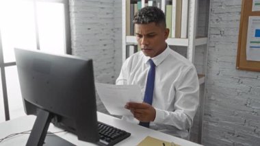 Ofisteki genç adam yoğun bir şekilde masa başında doküman okuyor, hayal kırıklığı sergiliyor, modern iç mekanla, odaklanmış profesyonel bir ortam öneriyor..