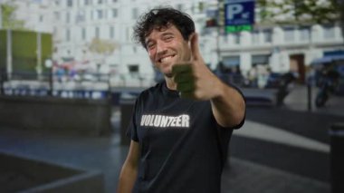 Açık havada gülümseyen gönüllü gömlekli bir adam şehir caddesinde baş parmak işareti yapıyor..