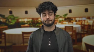 Genç adam sessiz restoranda meditasyon yapıyor, gözleri kapalı, masalar ve bitkilerle çevrili, huzurlu bir ortam yaratıyor..