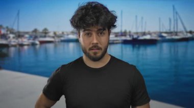 Güneşli bir günde arka planda tekne ve gemilerle dolu canlı bir limanda duran sakallı genç bir adam..