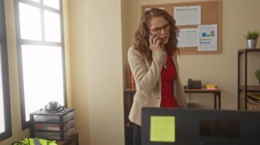 Ofiste bilgisayar masası ve iç dekorasyonla telefonda konuşan, iş iletişimi ve işyeri dinamiğine odaklı profesyonel çalışma ortamı sergileyen bir kadın..