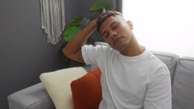 Genç İspanyol bir adam, rahat bir oturma odasında rahat bir şekilde dinleniyor ve boynunu uzatıyor, modern dekorlu, günlük beyaz gömlek giyiyor, rahat ev ortamını ön plana çıkarıyor..