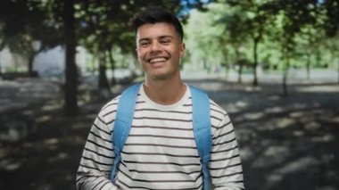 Mavi sırt çantalı genç adam, yemyeşil ağaçlarla çevrili güneşli bir açık hava parkında sıcak bir şekilde gülümserken başparmağını kaldırıyor..