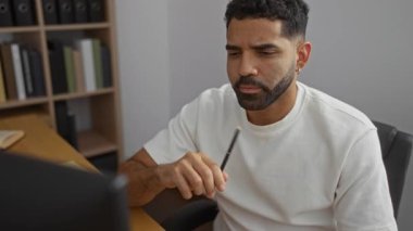 Yakışıklı, genç, İspanyol bir adam bir ofiste arka planda kitap rafları olan bir kalemle dalgın dalgın oturuyor.