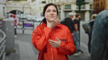 Turuncu hapishane tulumu giymiş bir kadın, rahat hissetmiyor, arka planda insanlarla dolu bir caddede duruyor, açık hava manzarası sergiliyor..