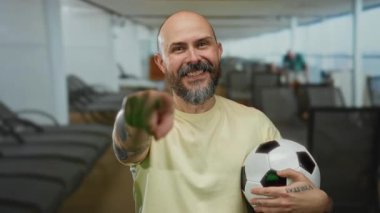 Futbol topu tutan ve parlak gökyüzünün altında bir yolcu gemisinde duran sakallı neşeli kel adam.