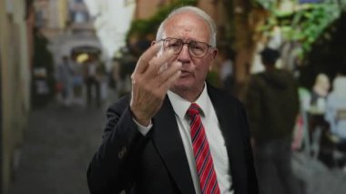 Takım elbiseli kıdemli işadamı yoğun bir caddede öfkeyle el kol hareketi yapıyor. Yüzü bulanık yayalar ve açık hava manzarasıyla çevrili..