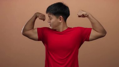 Genç Çinli bir adam kaslarını kırmızı bir gömlekle kahverengi bir arka plana yaslar ve gururlu, atletik bir duruşla gücünü ve güvenini gösterir..