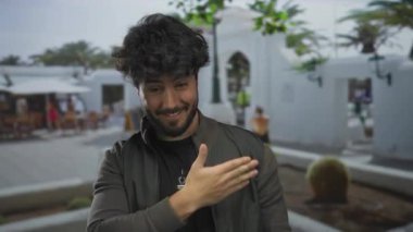 Güneşli bir günde arka planda insanlar ve palmiyelerle gülümseyen İspanyol genç bir adam. Günlük ceket ve siyah gömlek giymiş, açık havada canlı bir atmosfer yakalıyor..