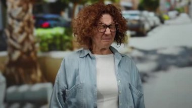 Orta yaşlı İspanyol bir kadın, bir şehir caddesinde elini yüzünü kapatıyor, gözlük ve kot gömlek takıyor, şehir dışında bulanık bir arka planda duygularını yansıtıyor..