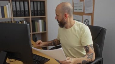 Sakallı ve dövmeli İspanyol bir adam ofiste oturuyor. Gözlükleri ayarlıyor ve bilgisayarlı belgeler okuyor. Etrafı klasörlerle çevrili ve odaklanmış bir iş ortamı yaratıyor..