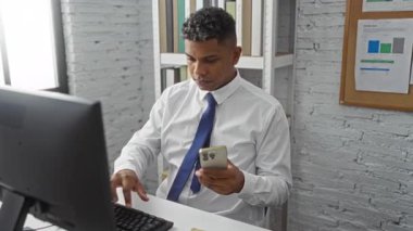 Ofisinde bilgisayar başında çalışırken arka planında modern bir iç mekan ve kitaplık olan İspanyol bir adam akıllı telefon kullanıyor..