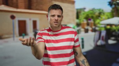 Kırmızı çizgili gömlekli genç adam, şehir sokağında kolunda dövmeleri olan etkileyici hareketler yapıyor.