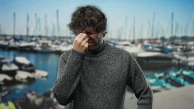 Genç adam deniz kenarındaki bir limanda gündüz vakti arka planda teknelerle stresli görünüyor..