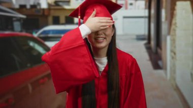 Kırmızı mezuniyet kıyafetli kadın açık havada akademik başarıyı kutlayan şehir caddesinde neşe içinde gözlerini kapatıyor..