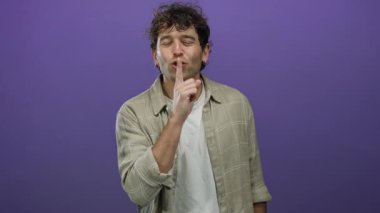 Kıvırcık saçlı İspanyol bir adam mor arka plana karşı sessiz jest yapıyor. Günlük gömlek ve küpeler takıyor. Gizlilik ya da sessizlik ima ediyor.