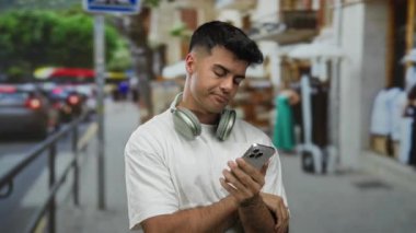 Kulaklıklı genç adam gündüz vakti şehir caddesinde akıllı telefonu kontrol ediyor..