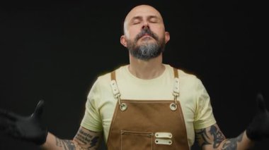 Sakallı ve dövmeli bir adam siyah arka plana karşı meditasyonla poz verir kahverengi önlük ve siyah eldivenler giyer..