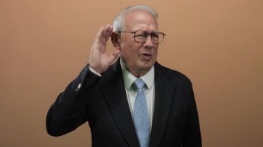 Gözlüklü kıdemli bir adam dikkatle dinliyor, kulaktan kulağa, tenha kahverengi bir arkaplan önünde, merak ve anlayışı vurguluyor.