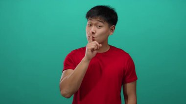 Genç adam, kırmızı gömlekli, izole edilmiş yeşil arkaplan karşısında sessiz bir şekilde el kol hareketi yapıyor.