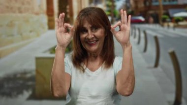 Üst düzey bir kadın iki eliyle iyi bir jest yapıyor, güneşli bir şehir caddesinde neşeyle gülümsüyor, şehir merkezindeki mutluluk ve pozitifliği yansıtıyor..