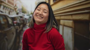 Canlı kırmızı bir süveter içinde gülümseyen genç bir kadın açık bir günde kalabalık bir şehir caddesinde park etmiş arabalar ve şehir mimarisi ile çevrili duruyor..