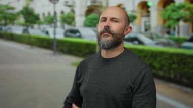 Kel ve sakallı bir adam, şehir sokaklarında bıyıklarını düzeltiyor, kendine güvenen bir stil sergiliyor ve modern bir erkek, manzaralı bir kasaba ortamında bakımını yapıyor..