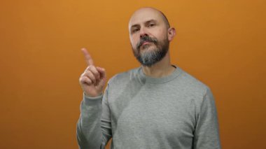 Kel ve sakallı bir adam, elini turuncu arka planda gezdiriyor, gri bir gömlek giyiyor, iletişim kuruyor ve ifade ediyor..