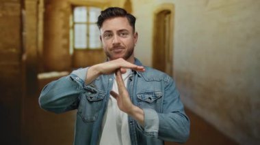 Kot ceketli genç adam eski bir üniversite binasında sıcak aydınlatma, rahatlama ve duraklamayı sembolize eden bir jest yapıyor..