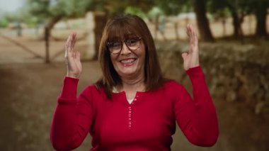 İspanyol son sınıf öğrencisi, gülümseyen ve el kol hareketi yapan, açık hava parkında kaldırılan kırmızı bir kazak giyen, ağaçlarla çevrili mutluluk ve pozitifliği ifade eden bir kadın..