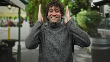 Genç bir adam gülümsüyor ve bir çerçeve jest yapıyor, ellerini şehir sokaklarında, neşeli bir ifade ve günlük bir stil ile canlı bir anı yakalıyor..